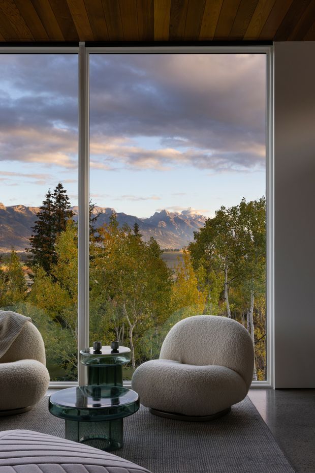 A refined living room with expansive floor-to-ceiling glazing framing a dramatic mountain and forest landscape. The interior features sculptural cream boucle lounge chairs, a low rounded glass coffee table, a textured grey rug, a partially visible upholstered sofa, and a dark timber-lined ceiling. The atmosphere is calm, luxurious, and nature-connected.