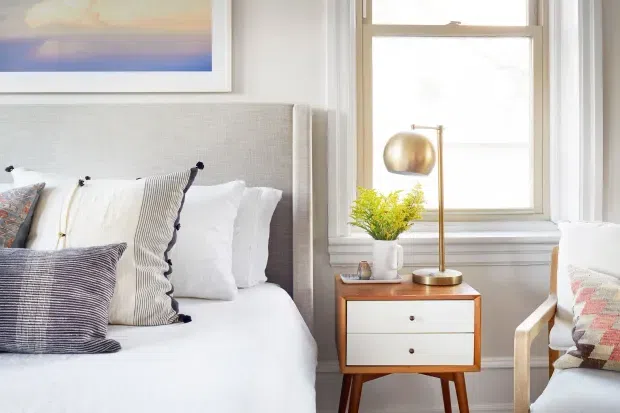 Bright, refined bedroom vignette featuring a neatly made bed with a textured upholstered headboard, layered white and patterned pillows, and a mid-century-style timber bedside table with white drawers. A brass globe lamp, small plant, framed artwork, and large white-trimmed window add warmth and natural light.