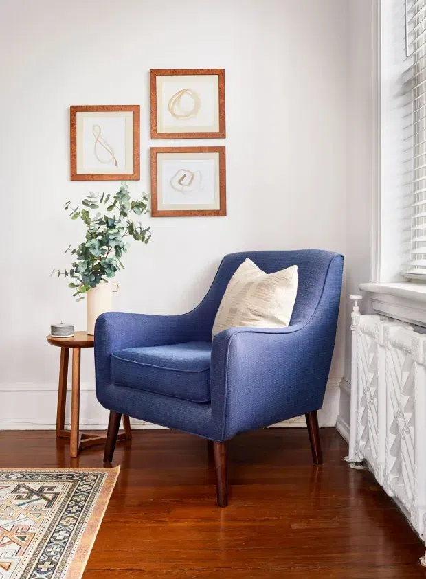 A bright, compact sitting corner with a deep blue upholstered armchair, cream accent cushion, small wooden side table, tall vase of leafy branches, framed abstract artwork, and a patterned rug. The space has white walls, polished dark timber flooring, traditional white window trim, and a decorative radiator cover beneath a large window with white blinds.