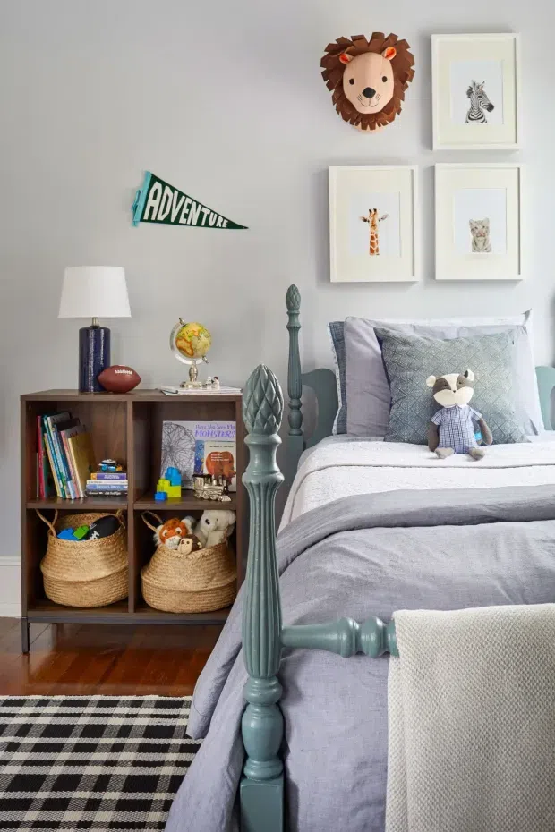 A cozy, playful children's bedroom with a grey upholstered or painted wall, a vintage-style dark teal spindle bed, layered grey and blue bedding, and a black-and-white checkered rug over timber flooring. A dark wood open bookshelf holds books, toys, baskets, a lamp, and decorative objects. The wall features framed animal illustrations, a lion wall sculpture, and an 'Adventure' pennant. Soft natural light and muted colours create a warm, inviting atmosphere.