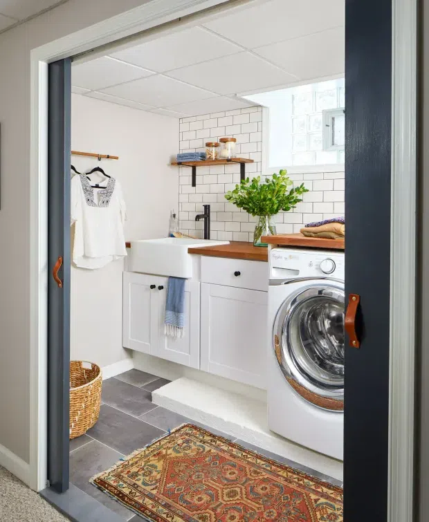 Bright, compact laundry room viewed through a doorway, featuring white shaker-style cabinetry, a deep white utility sink, wood benchtop, white subway-tile splashback, front-loading washing machine, open shelving, hanging clothes, and a patterned runner rug. A glass-block window provides natural light, while dark blue-grey door trim and leather handles add contrast.