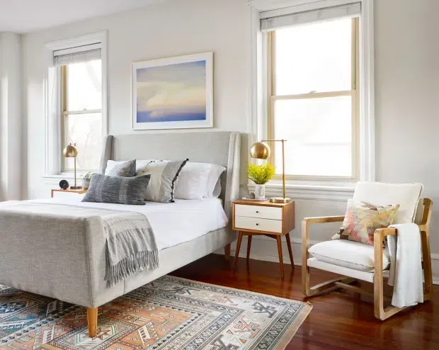 Bright, inviting bedroom with a light upholstered bed positioned between two tall windows, white bedding, layered neutral and patterned pillows, a small mid-century-style bedside table with brass lamp and plant, and a wooden-framed armchair. Dark timber flooring is partially covered by a large patterned rug, while pale walls and framed artwork create an airy, refined interior.