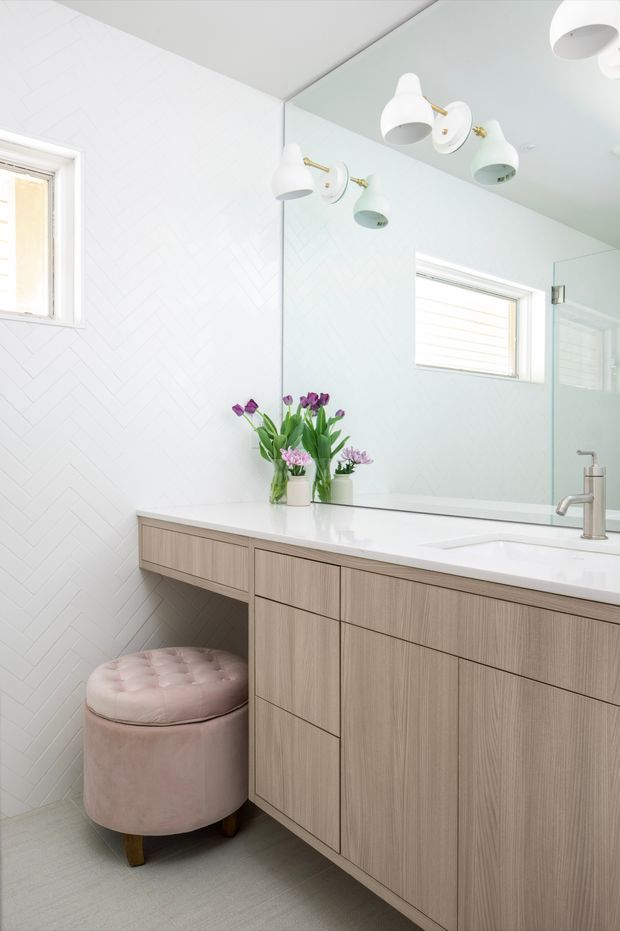 Bright contemporary bathroom vanity with pale wood-grain cabinetry, a white engineered stone benchtop, integrated basin, wall-to-wall mirror, white herringbone wall tile, and brushed metal tapware. A small blush upholstered stool and flowers add soft colour.