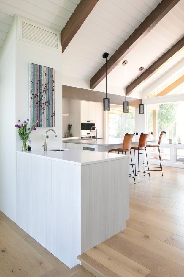 Bright open-plan kitchen with a large white island, integrated sink and cabinetry, pale timber flooring, white shiplap-style ceiling, exposed dark timber beams, pendant lights, and a breakfast bar with leather stools. Large windows bring in abundant natural light, and a colourful vertical artwork adds a decorative accent.