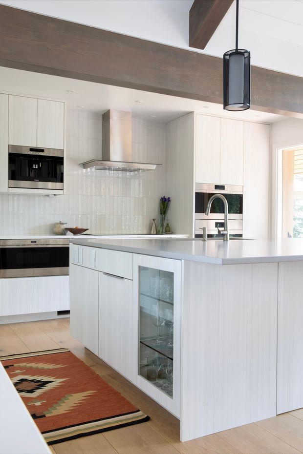 Bright contemporary kitchen with a large white island, integrated appliances, pale vertical-grain cabinetry, a light stone benchtop, and a glossy white tiled splashback. A substantial dark timber ceiling beam and matching hanging pendant add contrast, while a patterned rug introduces warm terracotta and earthy tones.