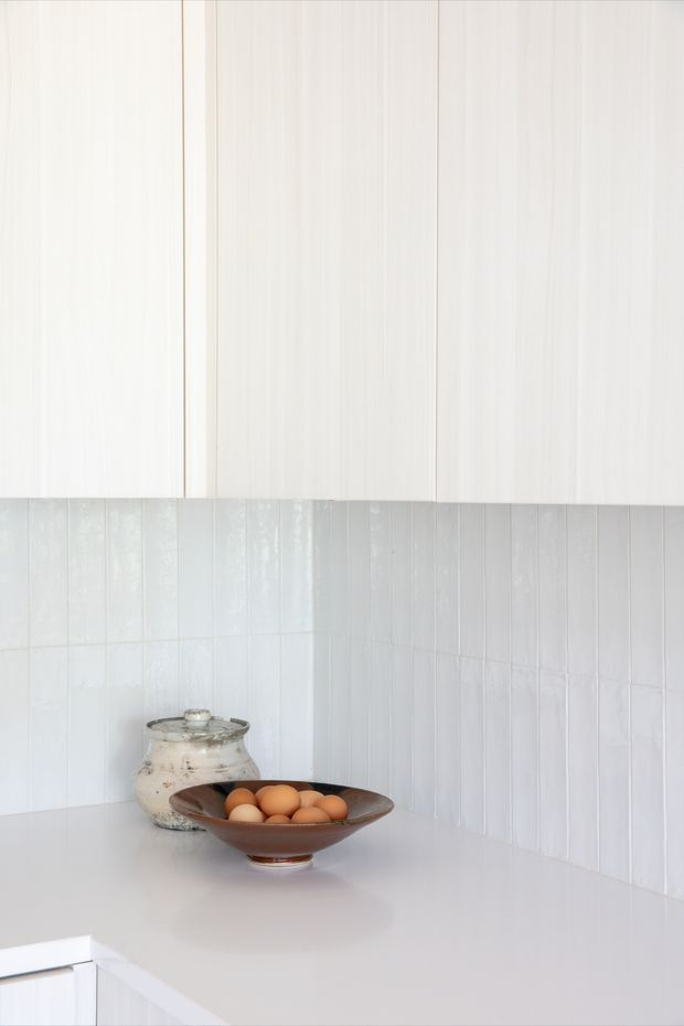 A close-up of a bright, minimalist kitchen corner with pale vertically grained cabinetry, glossy white vertical wall tiles, and a smooth white benchtop. A ceramic vessel and wooden bowl with eggs provide subtle warm accents.