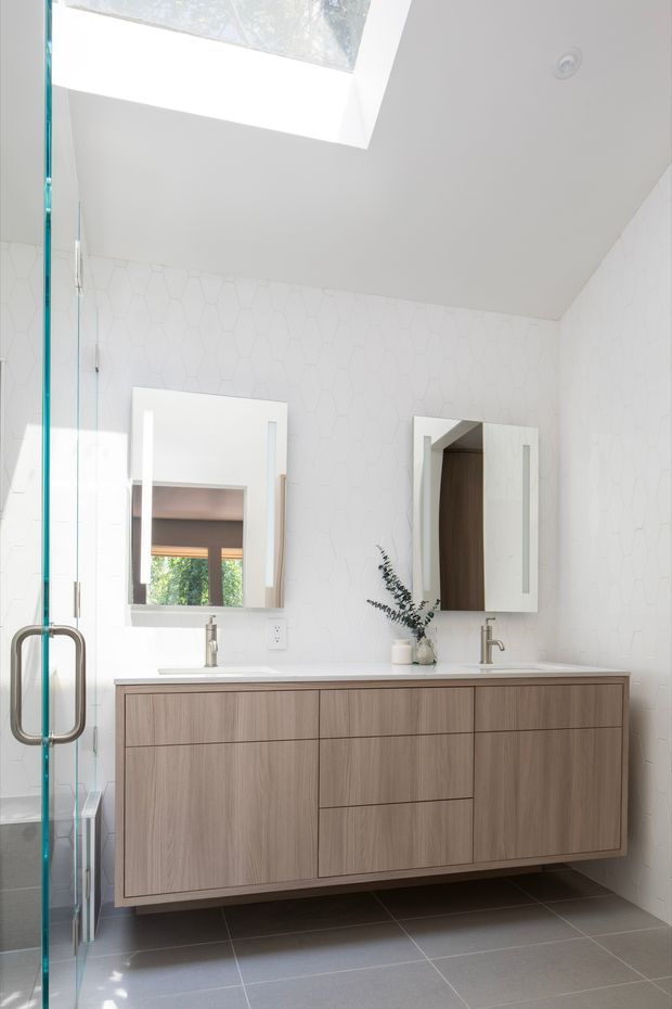 Bright contemporary bathroom with a floating double vanity in light wood cabinetry, white countertop, twin basins and brushed metal faucets. White geometric wall tiles, dark grey floor tiles, frameless glass shower enclosure, mirrored cabinets and a large skylight create a clean, airy space.