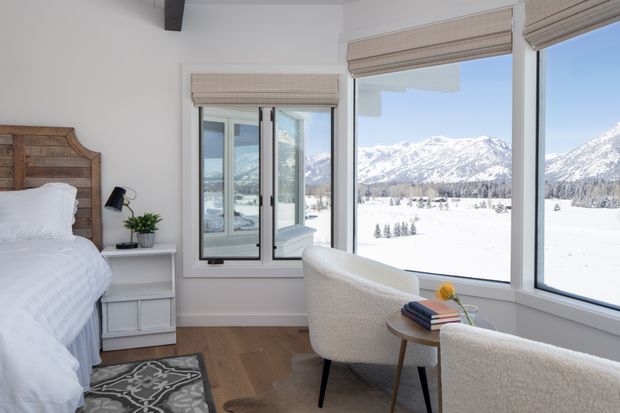 Bright bedroom with a rustic timber headboard, white bedding, light wood flooring, and two upholstered lounge chairs beside expansive corner windows. The glazing frames a dramatic snow-covered mountain landscape, while woven roller blinds and a small round side table add warmth and texture.