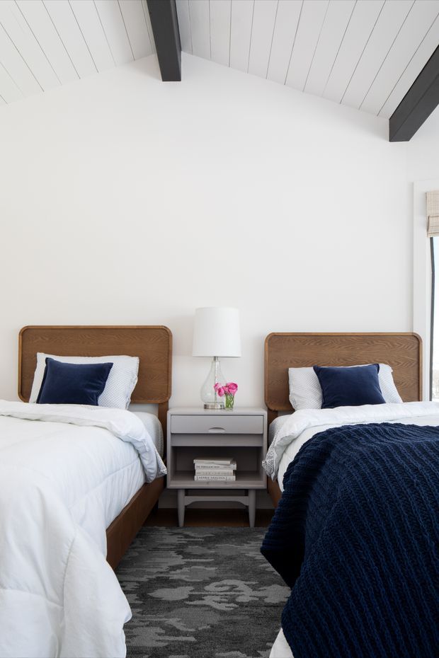 Bright, compact twin bedroom with two beds positioned around a shared grey bedside table. The room has white walls and a white slatted vaulted ceiling with prominent dark exposed beams. Warm timber bedheads, crisp white bedding, navy cushions and a navy knitted throw create a clean coastal-inspired look. A grey patterned rug, table lamp and small floral arrangement complete the space.