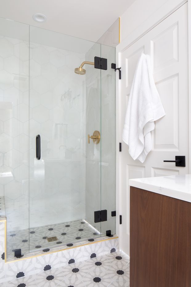 Bright, compact bathroom featuring a frameless glass walk-in shower with black hardware, brass shower fixtures, white hexagonal wall tile, and patterned encaustic-style floor tiles extending into the room. A white paneled door with a towel, black lever hardware, and a warm dark-wood vanity with a white benchtop are visible.