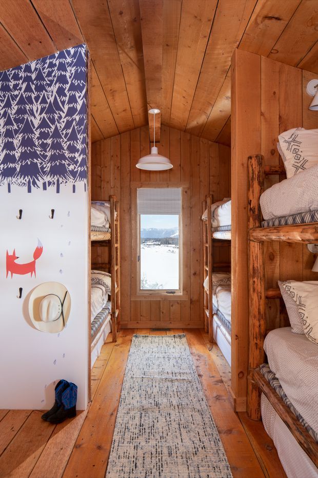 A compact rustic bunk room with timber wall and ceiling panelling, built-in log-framed bunk beds lining both sides, and a narrow central walkway. A patterned snowboard-like panel with hooks and hats stands at the left, while a small pendant light, window, and neutral runner complete the warm cabin interior. Snowy mountain scenery is visible through the rear window.
