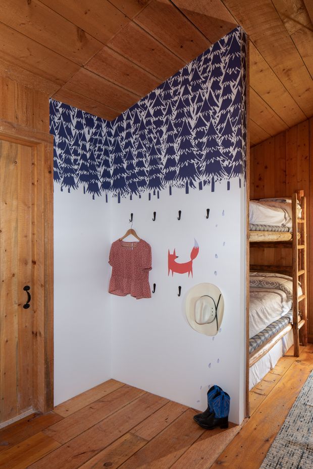 A compact, rustic timber-lined bunk room with a white feature wall decorated with a dark navy forest-tree pattern along the upper portion. The wall includes hooks holding a child's dress and a hat, while built-in wooden bunk beds with patterned bedding are visible to the right. Warm natural timber flooring, ceiling boards, and wall panelling create a cosy cabin-like atmosphere.