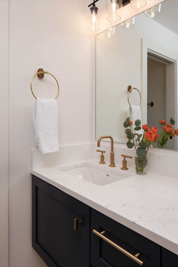 A refined bathroom vanity with a white engineered stone countertop, undermount sink, dark navy or black shaker-style cabinetry, and brushed brass faucet and hardware. A large wall mirror reflects the vanity and doorway, while a brass towel ring, contemporary glass pendant lighting, and a vase of flowers add decorative detail. The compact space has a warm, elegant finish.