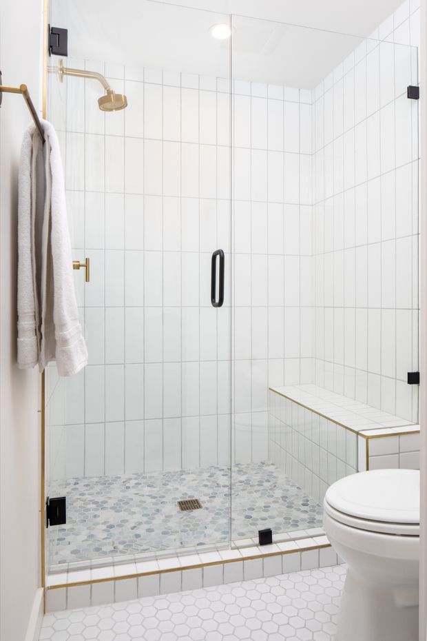 Bright contemporary bathroom featuring a large frameless glass walk-in shower with white grid wall tiles, blue-grey pebble mosaic shower flooring, a tiled built-in bench, brushed brass shower hardware, and a white hexagonal floor tile outside the shower. A white toilet and wall-mounted towel rail are visible.