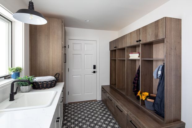 A bright, compact entryway and laundry drop zone with a white utility sink beneath a window on the left and a large built-in dark timber mudroom unit along the right wall. The joinery includes open cubbies, upper cabinets, drawers, hanging space, and black hardware. A white paneled entry door sits at the end, while a patterned dark floor adds contrast. The room has a practical, warm contemporary character with abundant natural light.