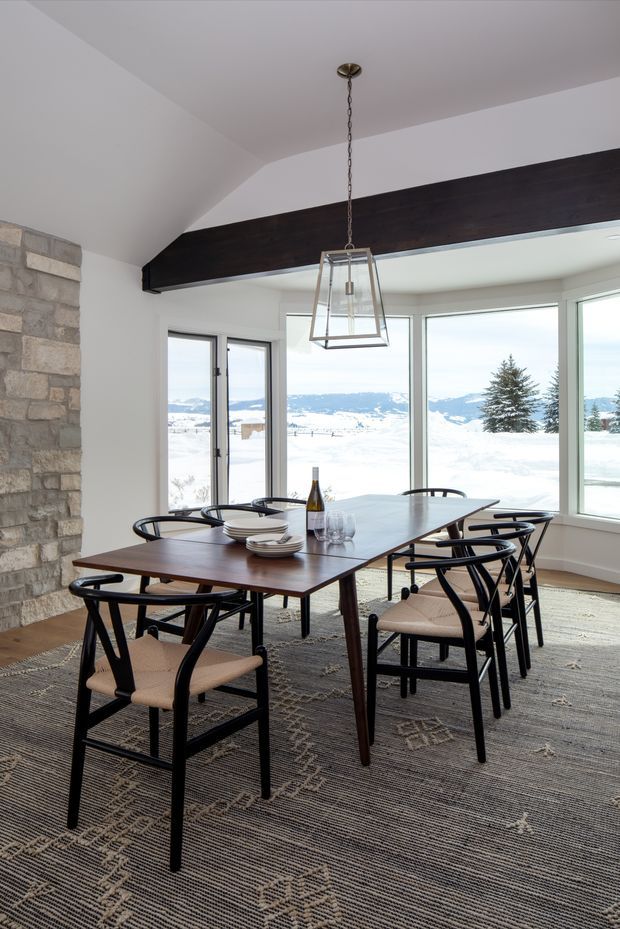 Bright dining room with a large dark timber dining table surrounded by black-framed upholstered chairs. A substantial exposed dark timber beam crosses the vaulted white ceiling, while a stone-clad wall and contemporary pendant light add texture. Full-height corner glazing overlooks a snowy mountainous landscape.