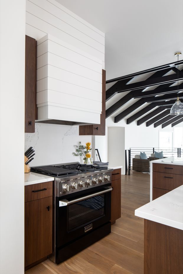 A contemporary open-plan kitchen featuring dark walnut cabinetry, white horizontal wall panelling, a white marble-look backsplash, and a large professional black range cooker with stainless controls. White island benchtops frame the foreground, while the adjoining living area has dark exposed ceiling beams, timber flooring, black-framed balustrading, and pendant lighting.