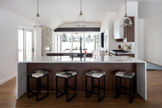 Bright open-plan kitchen viewed from the dining or living area, featuring a large white waterfall-edge island with dark timber cabinetry, four black-framed bar stools, pendant lights, white cabinetry, timber flooring, and a stone feature wall extending toward the glazed living area.