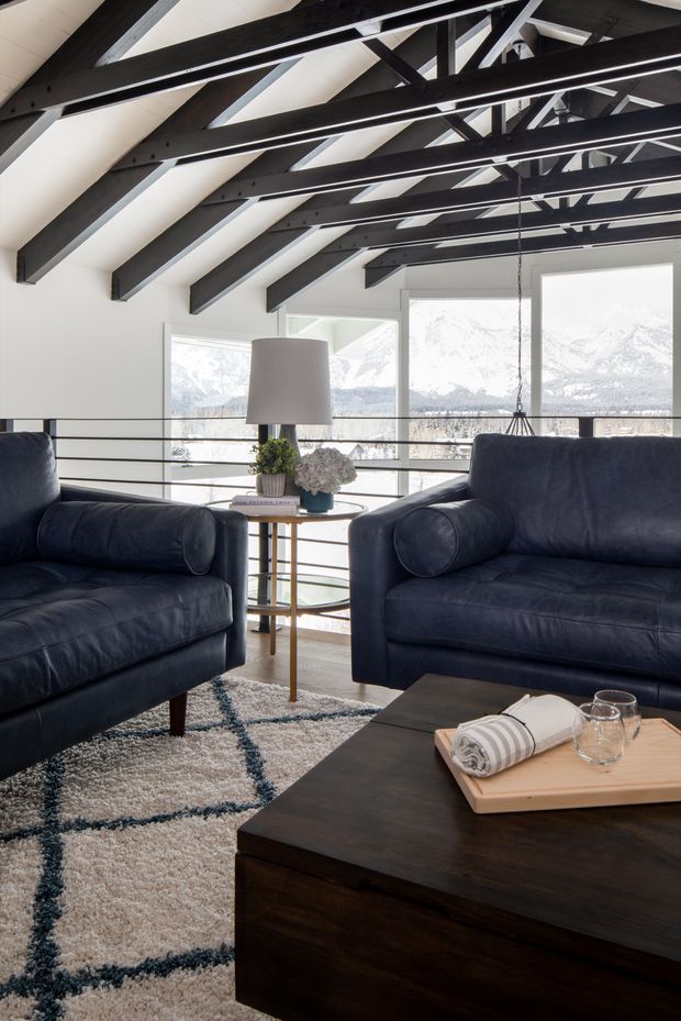 A sophisticated living room with two deep navy leather sofas facing a large dark wood coffee table. A textured light rug with dark geometric lines anchors the seating area. The room features dramatic exposed dark timber roof trusses and beams, white vaulted ceilings, pendant lighting, and expansive windows overlooking a snowy mountain landscape. A slim black metal balustrade separates the living area from the glazed edge, creating an airy alpine setting.