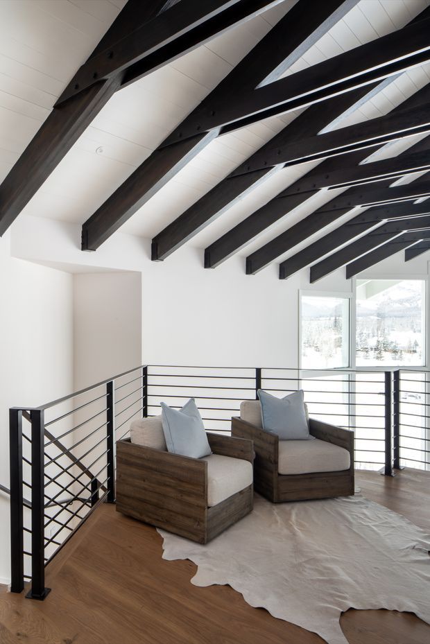 A bright loft-like sitting area beneath a sloped ceiling, featuring exposed dark timber beams, white panelled ceiling and walls, dark metal horizontal railings, warm timber flooring, and two rustic wood-framed lounge chairs with pale upholstered cushions. Large glazing reveals a snowy landscape outside.