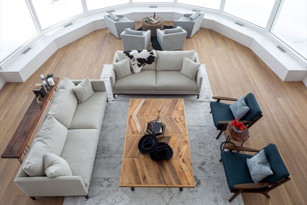Bright, spacious living room arranged around a large rustic timber coffee table, with two light upholstered sofas, navy accent lounge chairs, a grey area rug, and a separate seating arrangement near expansive angled windows. The room features light timber flooring, white built-in window surrounds, and abundant natural light.