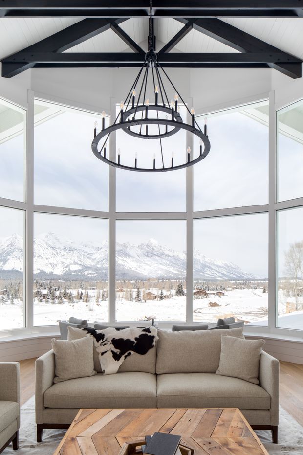 Bright living room with a large sectional-style sofa and rustic wood coffee table, set before expansive floor-to-ceiling corner glazing overlooking a snowy mountain landscape. The room features a vaulted white ceiling with exposed dark timber trusses and a dramatic black multi-ring chandelier.
