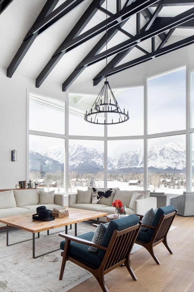 A bright, spacious living room with a vaulted white ceiling supported by exposed dark timber beams and trusses. Large floor-to-ceiling windows provide expansive mountain views. The room contains light upholstered sofas, a timber coffee table, blue upholstered timber lounge chairs, a large neutral rug, and a distinctive black circular chandelier. The interior combines a warm natural palette with dramatic structural detailing and alpine scenery.
