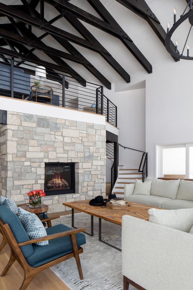 A bright, spacious living room with a large natural stone fireplace, exposed dark timber roof trusses, and a mezzanine level with black horizontal metal railings. The room includes a light upholstered sofa, blue mid-century-style armchairs, a substantial timber coffee table, a contemporary black chandelier, and a stairway leading to the upper level. White walls and high ceilings create an airy, refined atmosphere.