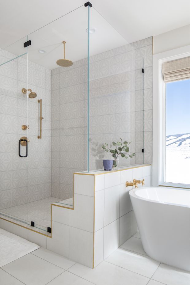 Bright contemporary bathroom featuring a walk-in glass shower with patterned wall tiles, brass shower fittings and tapware, a freestanding white bathtub, white tiled surfaces, and a small built-in tiled partition with a plant. A window provides views of a snowy landscape.