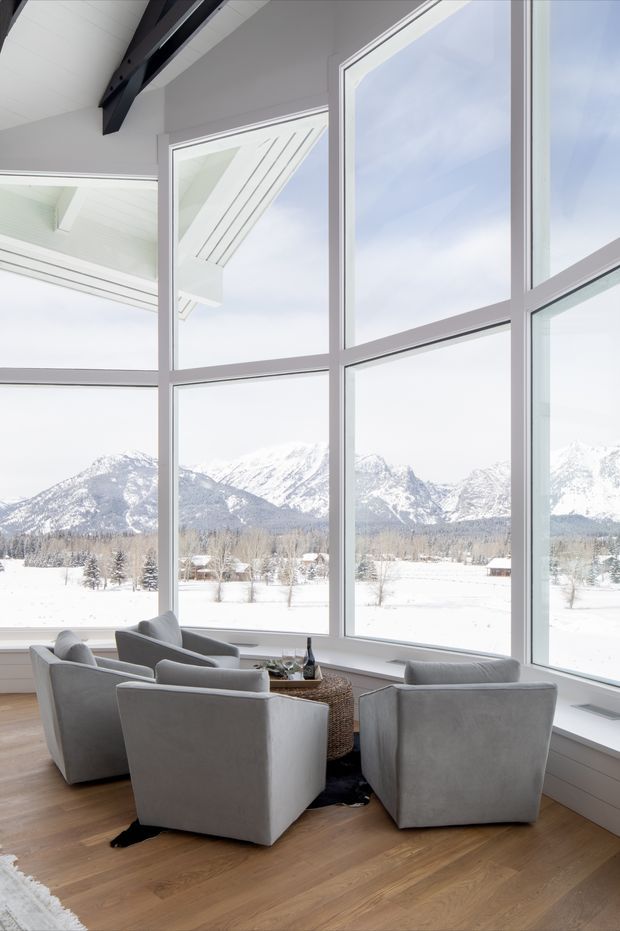 Bright double-height sitting area surrounded by expansive floor-to-ceiling glazing, with four grey upholstered armchairs arranged around a small round table. Pale timber flooring, white walls and ceiling, and dark exposed structural beams frame panoramic snowy mountain views.