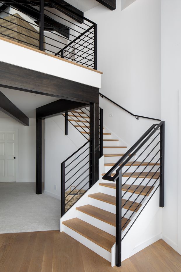 Bright contemporary interior focused on a staircase leading to an upper landing. The stair treads and surrounding flooring are warm timber, contrasted with crisp white walls and stair risers, while black metal balustrades, handrails, and exposed structural beams create a strong graphic element. An open mezzanine and adjacent carpeted room are visible in the background.