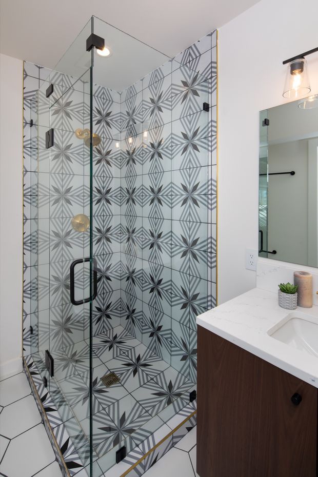 A contemporary bathroom featuring a walk-in glass shower enclosure with bold geometric patterned wall and floor tiles, black hardware, and brass-toned shower fixtures. A white engineered-stone vanity top with integrated sink and dark timber cabinetry sits beside a large mirror. The space is bright with a graphic, refined finish.