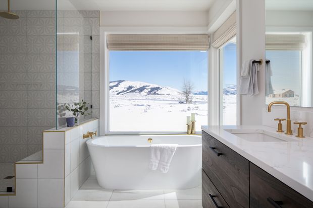 Bright, contemporary bathroom with a freestanding white bathtub positioned in front of large windows overlooking a snowy mountainous landscape. The room features patterned grey wall tiles, pale floor tiles, a timber-look vanity with a white stone benchtop, and brushed brass tapware and fixtures. A glass-enclosed shower is visible on the left, with roller blinds and a towel rail on the right.