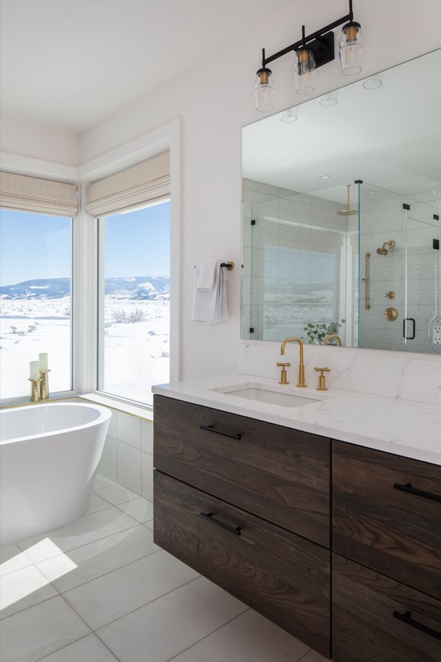 Bright, contemporary bathroom with a freestanding white bathtub positioned beside large corner windows overlooking a snowy landscape. The room features a dark wood floating vanity with black pulls, a white stone countertop and integrated basin, brass tapware, a large frameless mirror, and a glass-enclosed shower clad in patterned pale tiles. Black-and-brass vanity lighting and abundant natural light create a refined, warm atmosphere.