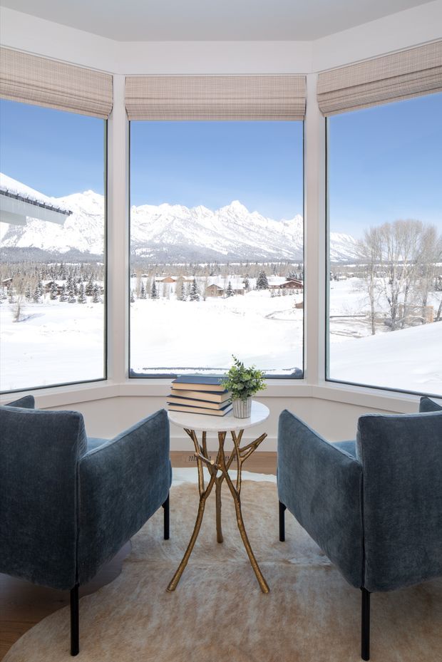 A bright, compact sitting nook with two deep blue upholstered lounge chairs facing a small round white table with a sculptural branch-like base. A stack of books and a potted plant sit on the table. Three tall corner windows provide expansive views of a snowy mountain landscape, with woven Roman blinds partially lowered. The room has pale walls, warm timber flooring, and a soft neutral rug.