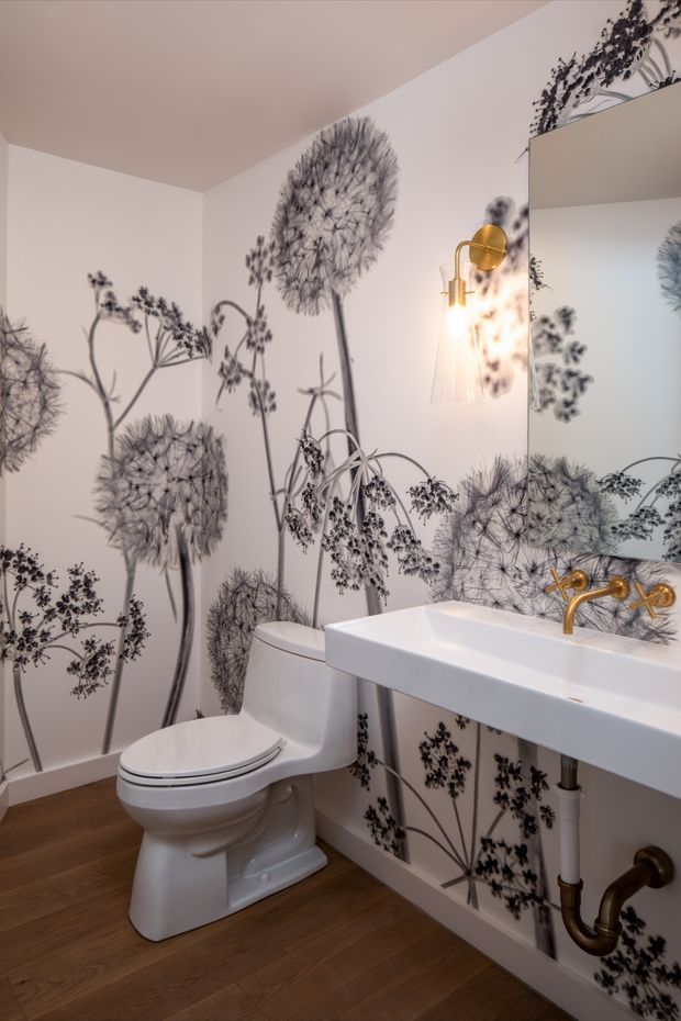 A compact powder room with a white toilet and wall-mounted rectangular basin, featuring striking black-and-white botanical wallpaper with oversized dandelion-like flowers. A large mirror, brass wall-mounted faucet, brass sconce, exposed plumbing, and warm timber flooring add decorative contrast.