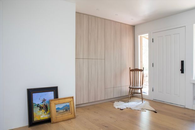 Bright, minimalist entryway with pale timber flooring, full-height light timber built-in cabinetry, white walls, and a white paneled front door with black hardware. A traditional wooden chair, small rug, and framed artwork add warmth to the otherwise clean, contemporary space.