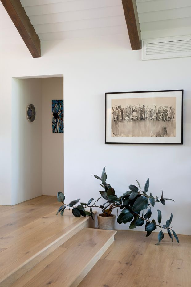 Bright interior stair landing with wide pale timber stairs and flooring, white walls, exposed dark timber ceiling beams, and a white-lined ceiling. A potted rubber plant sits beside the stairs, with framed artwork and decorative pieces visible near an open doorway. A high wall vent is visible near the ceiling.