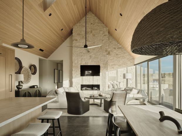Open-plan contemporary living, dining and kitchen interior with a dramatic vaulted timber-lined ceiling, exposed pale stone feature wall, fireplace and television. A large kitchen island occupies the foreground, while a dining table and upholstered seating sit beside full-height glazing. The palette is warm neutral with natural timber, stone, dark metal accents and abundant daylight.