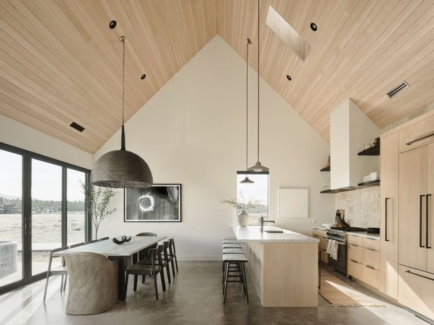 Open-plan kitchen and dining area with a dramatic vaulted ceiling clad in light timber boards. The room features a large pale-wood kitchen with island bench and stools, integrated cabinetry, open shelving, patterned backsplash, stainless appliances, pendant lights, a dark dining table with chairs, and full-height black-framed glazing. Polished concrete flooring and abundant daylight create a refined, warm contemporary interior.