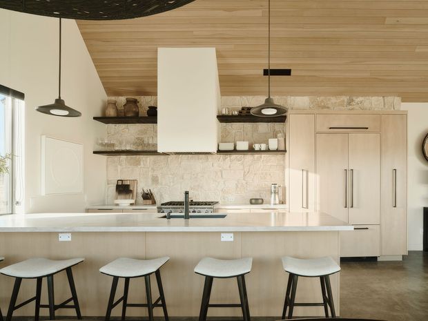 A spacious, warm-toned contemporary kitchen with a large pale island and four stools in the foreground. The room features light timber wall and ceiling panelling, a textured pale stone backsplash, integrated light-wood cabinetry and appliances, open shelving, a white rangehood, dark pendant lights, and a dark polished floor. A partial dining or living area is visible at the left edge.
