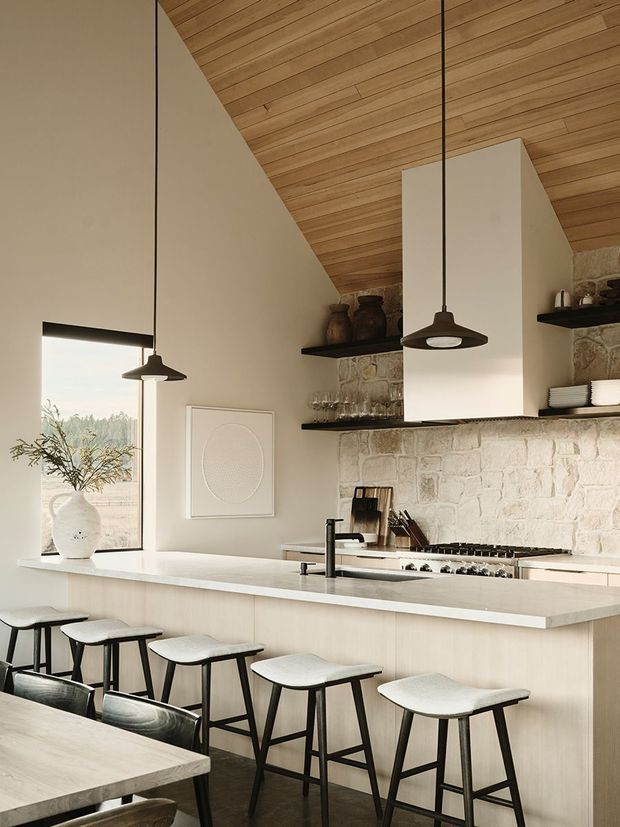 Warm, refined kitchen interior with a long pale island and integrated seating for several people. The room features a natural stone feature wall, white cabinetry and rangehood surround, open dark shelves with pottery and glassware, a timber-lined sloping ceiling, pendant lights, and large glazing overlooking a leafy landscape. A dining table is partially visible in the foreground.