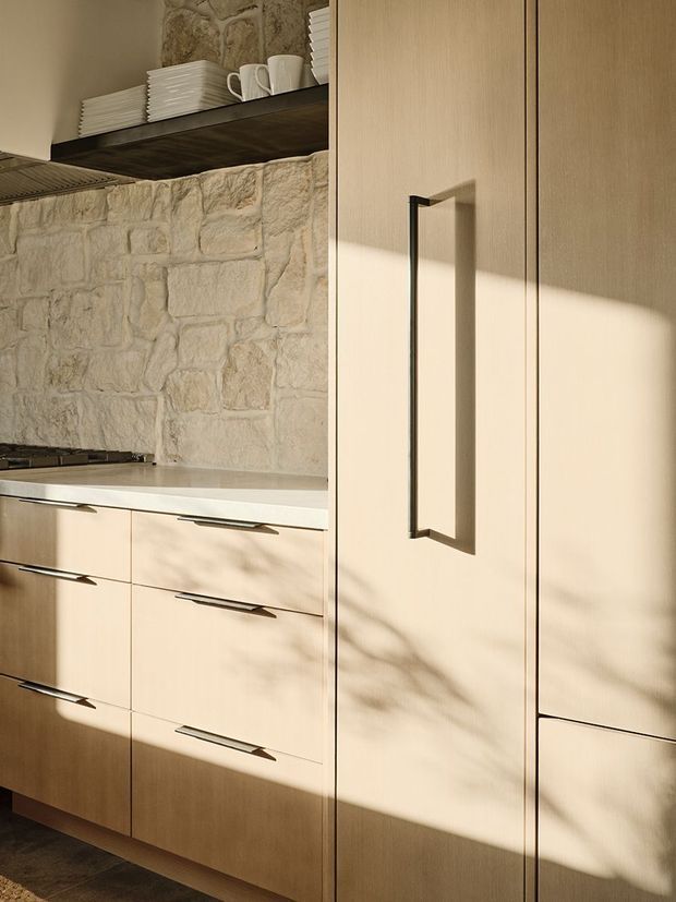 Close-up of a warm, refined kitchen featuring pale timber flat-panel cabinetry with long dark pull handles, a tall integrated storage unit, a light stone benchtop, and a rustic exposed stone wall backsplash. Open dark shelving displays neatly stacked white crockery. Strong sunlight creates soft organic shadows across the joinery.