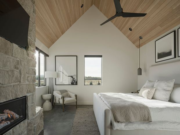 A bright, refined bedroom with a pitched ceiling clad in warm timber boards, white walls, and a substantial rough-stone fireplace wall with a built-in firebox. The room contains a neatly made upholstered bed, neutral textured bedding, a small upholstered lounge chair, floor lamp, framed monochrome artwork, and a ceiling fan. Tall narrow windows provide natural light and views beyond.