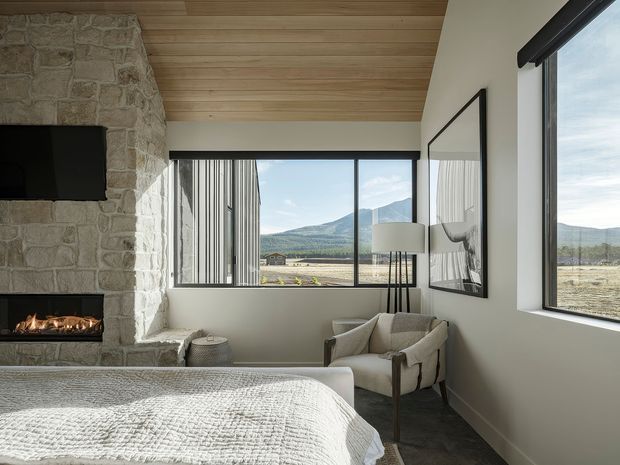 A refined bedroom with a large bed in the foreground, a stone fireplace and wall-mounted television on the left, and a comfortable armchair beside expansive windows. The room combines pale walls, a warm timber ceiling, dark flooring, and substantial glazing overlooking an open mountainous landscape. A large framed artwork, floor lamp, and textured bedding complete the calm, contemporary interior.