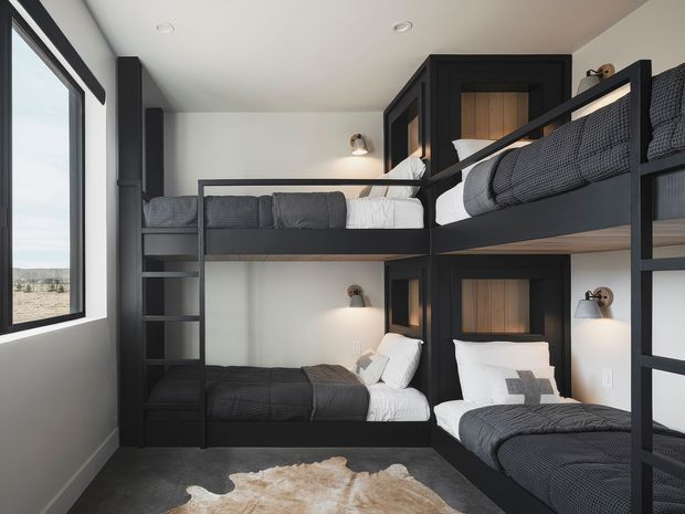 A compact bunk bedroom with four built-in sleeping berths arranged in two levels. The room features dark charcoal built-in bunk cabinetry and bed frames, warm timber-lined niches, integrated reading lights, dark bedding, white walls, and a large window overlooking an open landscape. A hide rug lies on the dark floor, creating a practical yet refined shared sleeping space.