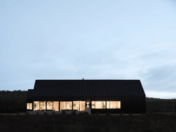 A dark, contemporary single-storey house set against a wooded rural landscape at dusk. The building has a simple barn-like form with a steep pitched gable roof, black vertically ribbed metal cladding, and a long band of warm illuminated windows along the front facade.