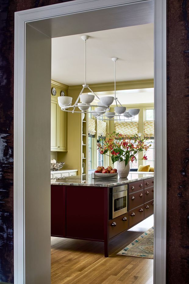 A richly detailed traditional kitchen viewed through a wide doorway. The room features olive-green cabinetry and decorative trim, a burgundy island with a dark stone benchtop, brown drawer cabinetry, integrated appliances, patterned window shades, wood flooring, and a contemporary multi-arm pendant light. A vase of flowers and fruit bowl sit on the island, with a patterned rug visible in the foreground.