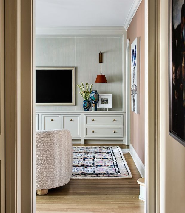 A refined living room viewed through a doorway, featuring a wall-mounted television, pale built-in cabinetry with brass knobs, a patterned area rug over timber flooring, a softly upholstered armchair, framed artwork, decorative ceramics, and a pendant wall light. The room has paneled or textured pale walls, substantial white ceiling trim, and a warm blush-toned side wall. The overall look is polished, traditional-modern, and layered with colour and pattern.
