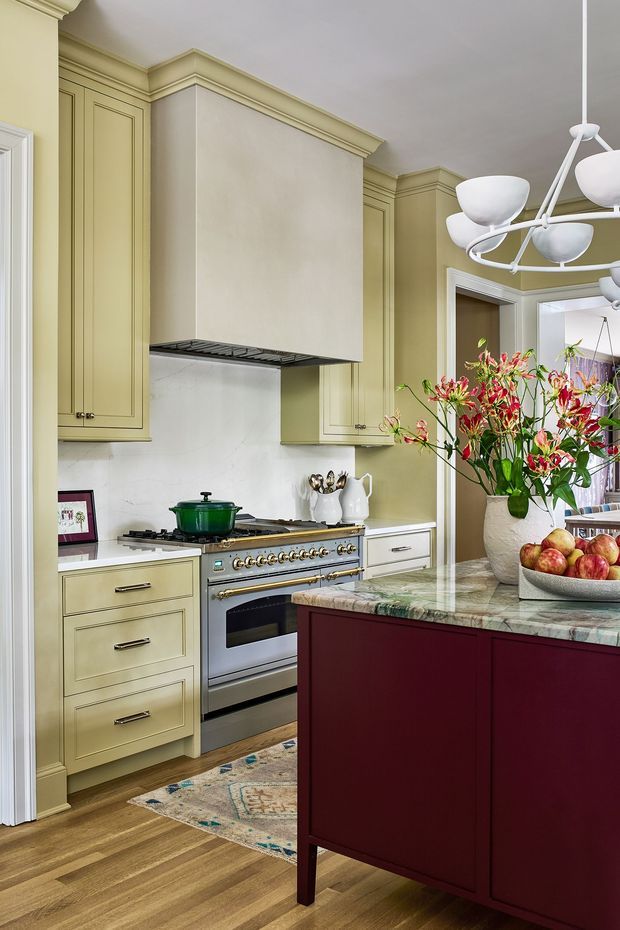 A refined traditional kitchen with pale sage-green raised-panel cabinetry, white marble-look splashback, a large burgundy island, and a veined stone benchtop. The room features timber flooring, classic crown moulding, brass hardware, a stainless steel range cooker, and a sculptural white multi-arm pendant light. A floral arrangement and bowl of fruit add colour and warmth.